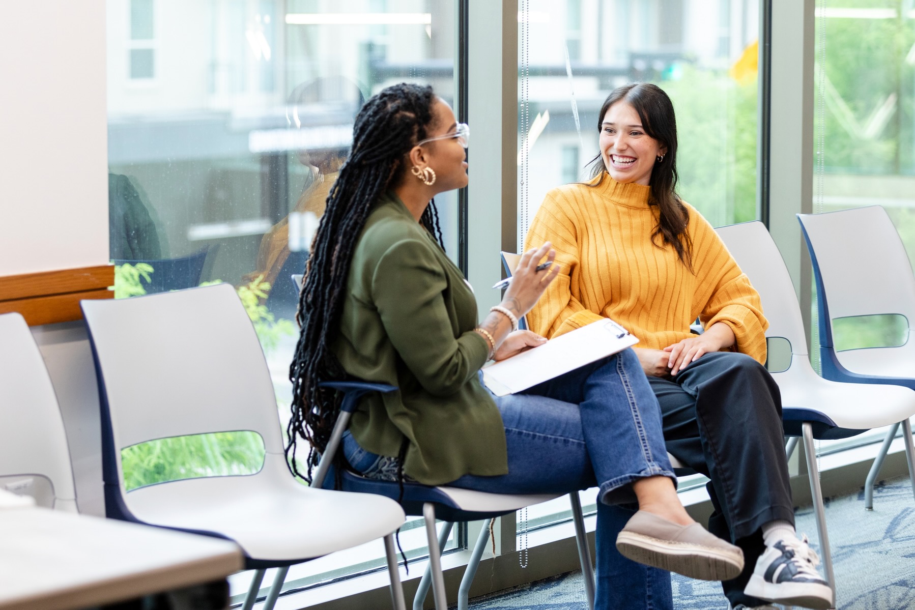 One woman with a clipboard on her lap is facing and talking to another seated woman who is smiling